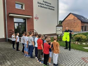 Policjant stoi przed wejściem do szkoły z grupą dzieci i prezentuje kamizelkę odblaskową.