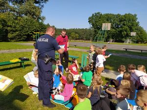 Policjant rozdaje dzieciom książeczki profilaktyczne na boisku przed szkołą