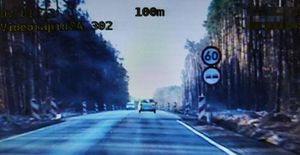 Droga widoczna przez kamerę radiowozu z wideorejestratorem. Przed radiowozem srebrny samochód. Przy drodze znak zakaz wyprzedzania i ograniczenie prędkości do 60 km/h.