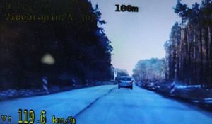 Droga widoczna przez kamerę radiowozu z wideorejestratorem. Przed radiowozem srebrny samochód. W prawym dolnym narożniku widoczna prędkość 119.6 km/h.