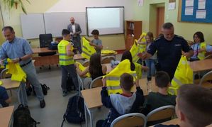 Policjant i przedstawiciel Kuratorium Oświaty w Opolu rozdają dzieciom żółte kamizelki odblaskowe.