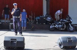 Policjant ruchu drogowego stoi na scenie amfiteatru. W prawej ręce trzyma mikrofon. Za nim w tle trzech mężczyzn, nagłośnienie i perkusja. Po prawej stronie zaparkowany czarny motocykl.