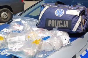 Na masce radiowozu leżą granatowa torba ze sprzętem do pierwszej pomocy z napisem POLICJA oraz zapakowane w folię zestawy do resuscytacji.