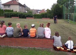 Dzieci siedzą na ławeczce patrzą w stronę boiska na którym policjant wykonuje pokaz tresury psa służbowego.