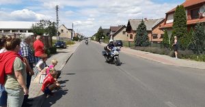 Skrzyżowanie. Po lewej stronie na chodniku stoją widzowie. Po jezdni jadą policjanci na szarych motocyklach jeden za drugim.