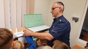 Policjant prezentuje jak wykonuje się daktyloskopię. Przy nim dzieci.