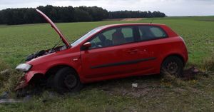 W tle pola uprawne i las. Na pierwszym planie widać lewy bok czerwonego samochodu osobowego z podniesioną pokrywą silnika. Samochód ma uszkodzony przód.