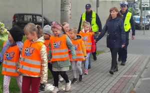 Dzieci spacerują po chodniku. Idą parami. Mają założone pomarańczowe kamizelki odblaskowe. Za nimi idzie opiekunka oraz dwóch policjantów w kamizelkach odblaskowych.