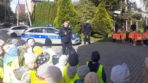 Policjant stoi na placu przedszkolnym i prowadzi pogadankę. Obok niego radiowóz. W tle drugi policjant. Na pierwszym planie przedszkolaki w kamizelkach odblaskowych. Dzieci stoją tyłem do kamery.
