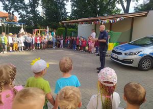 Policjant prowadzi pogadankę z przedszkolakami na placu przedszkolnym. Obok niego stoi radiowóz. Dzieci stoją w dużym kręgu.