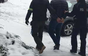 Policjant i policjantka prowadzą zatrzymanego na zewnątrz. Na ziemi lezy śnieg. Zatrzymany ma ręce z tyłu spięte kajdankami.
