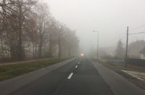 Jezdnia, w oddali widać światła nadjeżdżającego pojazdu, przejrzystość powietrza zmniejszona przez mgłę.
