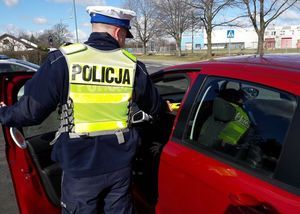 Policjant ruchu drogowego stoi w otwartych drzwiach czerwonego samochodu osobowego. W ręce trzyma urządzenie do badania stanu trzeźwości.