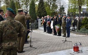 Na pierwszym planie po lewej stronie  widoczny Żołnierz Wojska Polskiego stojący przy Pomniku, w drugim planie stoją komendanci i dowódcy służb. W tle mogiły i drzewa.