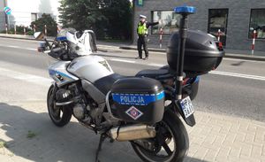 Na pierwszym planie policyjny motocykl. W tle policjant ruchu drogowego w kamizelce odblaskowej kieruje ruchem na drodze.