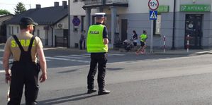 Policjant ruchu drogowego stoi na skrzyżowaniu. Obok niego stoi mężczyzna z żółtym napisem n a koszulce STRAŻ. Przed nimi wybiega ze skrzyżowania kobieta w seledynowej koszulce.
