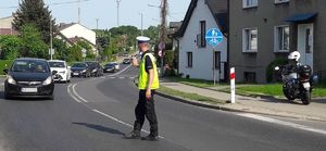 Policjant na ruchu drogowego stoi na skrzyżowaniu i zatrzymuje kolumnę samochodów. W tle zaparkowany policyjny motocykl.