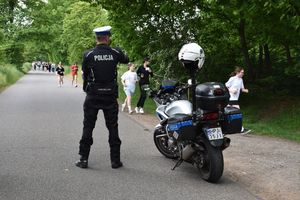 Policjant stoi obok motocykla. Przed nim biegnące dziewczęta.