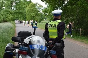 Policjant stoi obok motocykla i obserwuje grupę biegaczy, którzy biegną w jego kierunku.