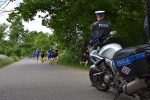 Policjant stoi przed zaparkowanym motocyklem. Obserwuje grupę biegaczy, którzy biegną w jego kierunku.
