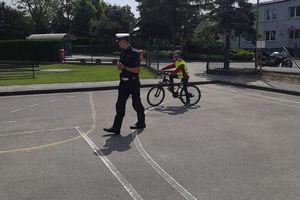 Policjant w białej czapce obserwuje ucznia kierującego rowerem, na asfaltowej nawierzchni widoczne namalowane znaki poziome.
