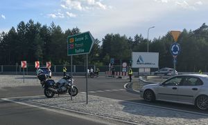 Rondo przy zjeździe na drogę ekspresową S11. Na drogach wlotowych stoją umundurowani policjanci zatrzymując ruch samochodów. Na środku ronda stoi policyjny motocykl.