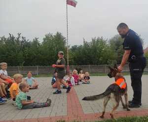 Plac zabaw. Dzieci siedzą na ziemi. Przed nimi stoi policjant, który trzyma za obrożę brązowego psa. Mały chłopiec niesie w ręku czerwoną małą piłkę. Pies patrzy w jego kierunku.