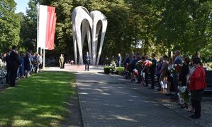Cmentarz komunalny w Oleśnie. W tle pomnik Lotników Polskich. Przy głównej alejce po prawej i lewej stronie stoją delegacje z wiązankami kwiatów.