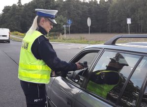Policjantka ruchu drogowego podaje urządzenie pomiarowe do badania stanu trzeźwości kierującemu srebrnym samochodem.