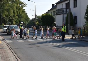 Dzieci przechodzą po przejściu dla pieszych mają uniesione w górze dłonie , policjanci stoją na jezdni, wstrzymują ruch pojazdów.