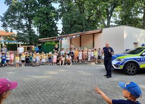 policjant stoi pośród przedszkolaków, obok niego radiowóz w nowych barwach ( srebrny , niebieski , żółty ).