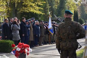 Na pierwszym planie po prawej stronie stojący Żołnierz Wojska Polskiego przy pomniku Lotników Polskich. W oddali po lewej stronie przedstawiciele służb mundurowych z uniesionymi prawymi dłońmi przy głowach.
