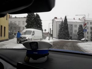 Widok przez przednią szybę radiowozu na ulicę Solny Rynek w Oleśnie. Na podszybiu leży czapka policjanta ruchu drogowego z białym pokrowcem z na niebieskim otoku jeden poziomy słupek. W tle po prawej stronie jedni mężczyzna w niebieskiej kurtce odśnieża szybę białego samochodu dostawczego.