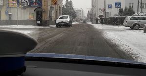 Widok przez przednią szybę radiowozu na ulicę Kościuszki w Oleśnie. Na podszybiu leży czapka policjanta ruchu drogowego z białym pokrowcem. W biały samochód jadący z przeciwnego kierunku. Na chodniku piesi. Na jezdni leży błoto pośniegowe. Chodniki i pobocze pokryte śniegiem.