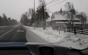 Widok przez przednią szybę radiowozu na ulicę Gorzowską w Oleśnie. Na podszybiu leży czapka policjanta ruchu drogowego z białym pokrowcem. Po lewej stronie kościółek św Anny pokryty śniegiem.  Na jezdni leży błoto pośniegowe. Pobocze pokryte śniegiem.