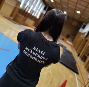 Hala sportowa zespołu szkół. Tyłem do obiektywu stoi uczennica w czarnej koszulce z widocznym na plecach napisem klasa mundurowa Dobrodzień.