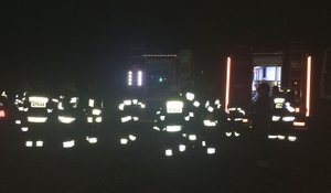 Strażacy z Ochotniczej Straży Pożarnej stoją przy wozach bojowych na miejscu zbiórki. W całkowitej ciemności widoczne elementy odblaskowe i napisy STRAŻ.