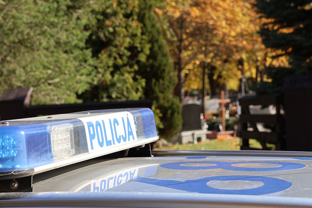 Zdjęcie przedstawia napis policja na dachu radiowozu w tle cmentarz