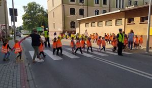 Dzieci w odblaskowych kamizelkach przechodzą przez jezdnię na przejściu dla pieszych. Policjanci w kamizelkach odblaskowych zatrzymują ruch podczas ich przejścia.