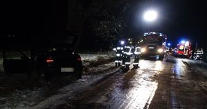 Zmrok. Ciemny samochód osobowy stoi na lewym poboczu drogi. Błyszcząca nawierzchnia drogi. W tle stoją ludzie w kamizelkach odblaskowych oraz ambulans i wóz bojowy straży pożarnej. Miejsce oświetla reflektor.