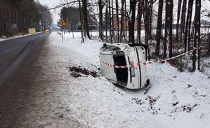 W rowie po prawej stronie drogi leży rozbity biały samochód. Rów i pobocze ośnieżone. Pojazd opasany taśmą ostrzegawczą w kolorze biało-czerwonym. Samochód leży na lewym boku.