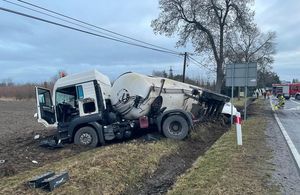 Uszkodzona cysterna leży na poboczu drogi. Kabina pojazdu skręcona, ustawiona prostopadle do drogi. Koła wryte w podłoże. Cysterna leży na skarpie rowu na prawym boku.W tle wóz bojowy straży pożarnej i strażak trzymający prądownicę.