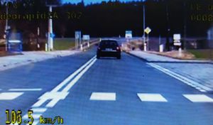Obraz widziany okiem kamery z radiowozu. Na jezdni przejście dla pieszych, a za nim ciemny samochód  widziany z tyłu. Po prawej stronie zatoczka autobusowa. W lewym dolnym narożniku kadru wyświetlono prędkość 106.5 km/h