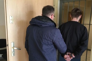 Nieumundurowany policjant trzyma zatrzymanego , który ma założone kajdanki na rękach trzymanych  z tyłu. Oboje stoją przed kratami do policyjnego aresztu.