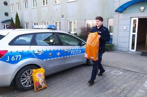 Umundurowany policjant niesie w rękach pomarańczowy worek z karmą. W tle oznakowany radiowóz, a obok stojący worek z karmą.