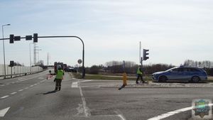 Miejsce wypadku drogowego. W tle widoczny wrak samochodu osobowego oraz wóz bojowy straży pożarnej. Policjantka prowadzi oględziny miejsca wypadku, obok niej stoi radiowóz. Strażak w ubraniu ochronnym i hełmie kieruję ruchem.