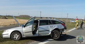 Srebrny samochód typu kombi widok lewej strony. Uszkodzony tył. Brak tylnego zderzaka. Otwarte przednie drzwi i podniesiona pokrywa silnika. W tle policjant w kamizelce odblaskowej oraz strażacy. Za nimi policyjny radiowóz.