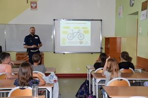 Policjant stoi przed siedzącymi w ławkach uczniami. Na tablicy wyświetlony slajd &quot;obowiązkowe wyposażenie roweru&quot;.