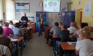 Policjant i przedstawiciel Wojewódzkiego Ośrodka Ruchu Drogowego z Opola stoją przed nauczycielami siedzącymi przy stolikach. Na  ścianie wyświetlony slajd &quot;Rowerowe wyzwanie&quot;.