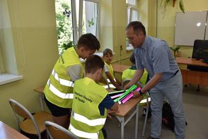 Mężczyzna rozdaję dzieciom siedzącym w ławkach odblaskowe opaski. Dzieci mają założone żółte kamizelki odblaskowe.